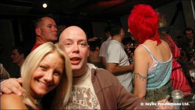 A blonde woman and a bald man in a brown hoodie smile at the camera in a crowd