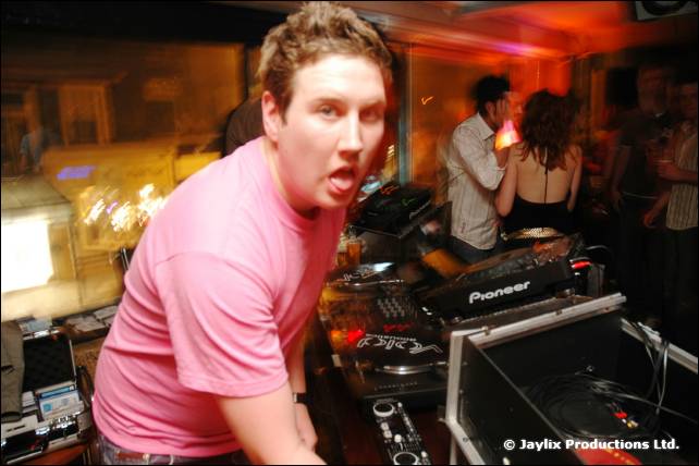 A man in a pink t-shirt sticks out his tongue beside Pioneer DJ decks