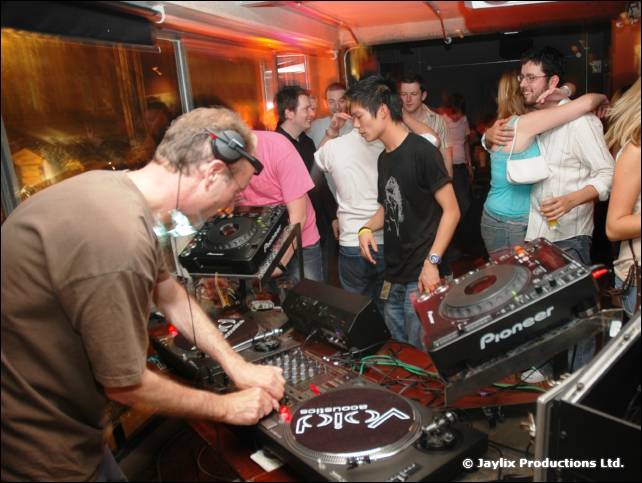A man in a brown t-shirt and headphones mixes at Pioneer decks while people stand behind him