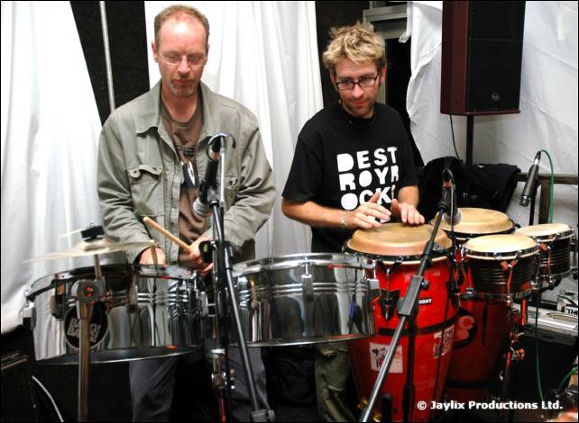 Two men play percussion, one in a green jacket and one in a black Destroy Rock t-shirt, inside a white tent