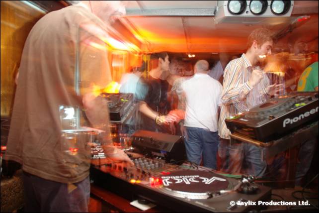 A blurred DJ in a brown top stands at Pioneer decks while a man in a striped shirt plays keyboards behind