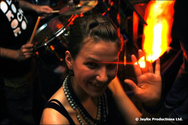 A smiling woman in a pearl necklace holds up a hand beside a bright flame, a drummer behind