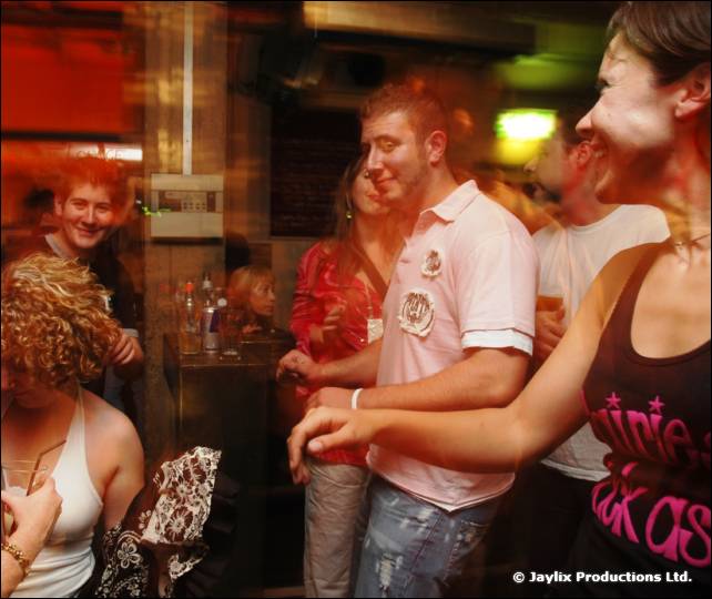 People dance in a bar, including a man in a pink polo shirt and a woman in a black top