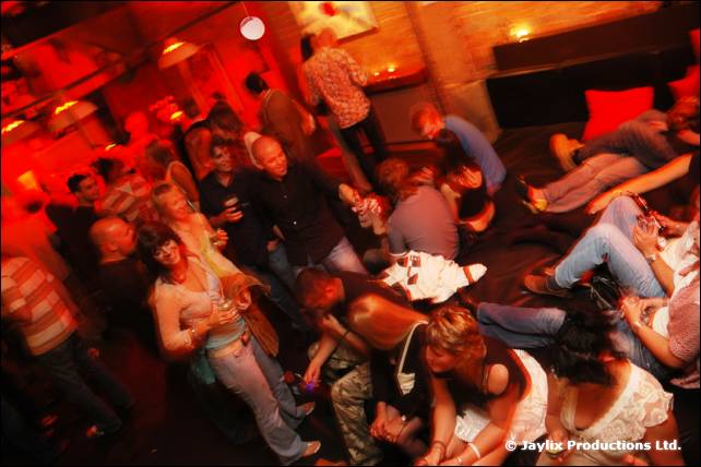 A crowd stands and sits in a red-lit bar, some lounging on cushions