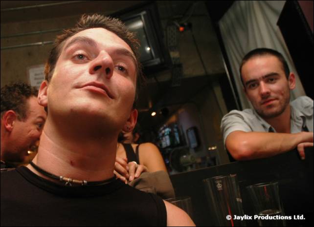 A man in a black vest looks down at the camera from a low angle, with another man behind at a table