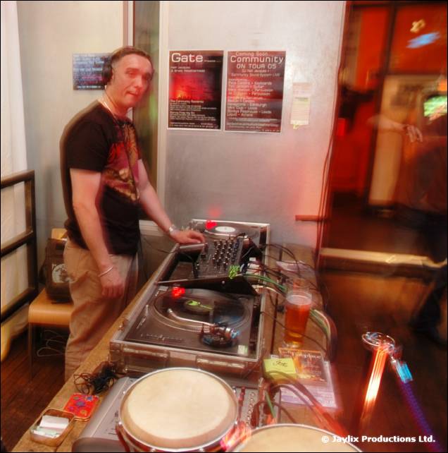 A DJ in a dark t-shirt and headphones stands at a turntable, with a bongo drum beside and posters behind