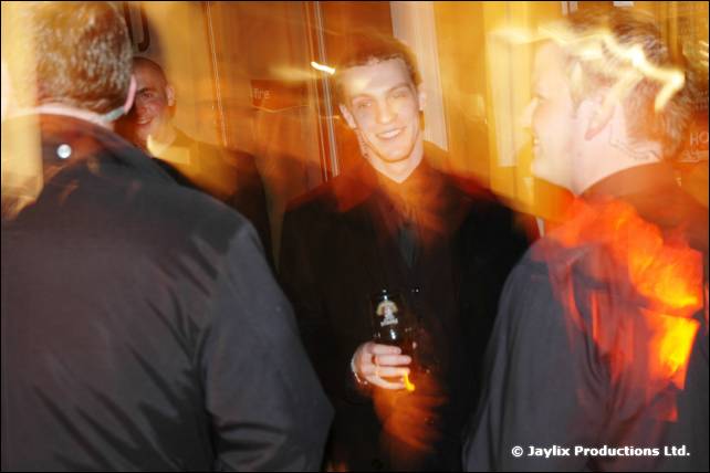 Three men stand close in a blurred crowd, the middle one smiling and holding a bottle