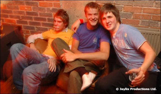 Three smiling young men sit close together on a sofa in front of a brick wall