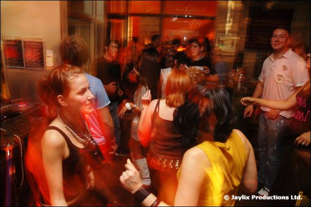 People dance in a busy bar, with a woman in a black top in the foreground and a man in a white shirt at right