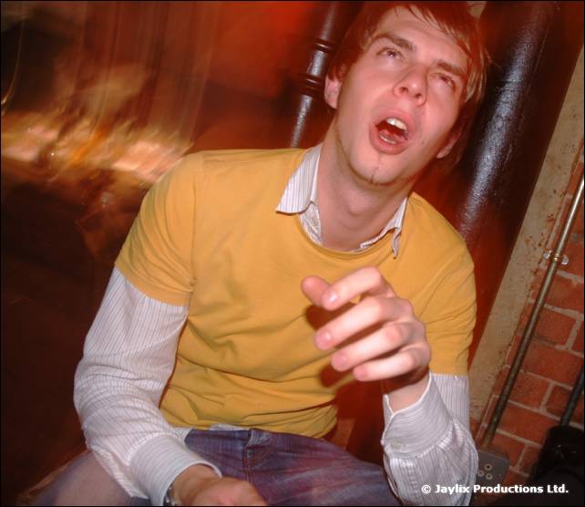 A person in a yellow top and striped shirt sits with mouth open, pointing, in a blurred club