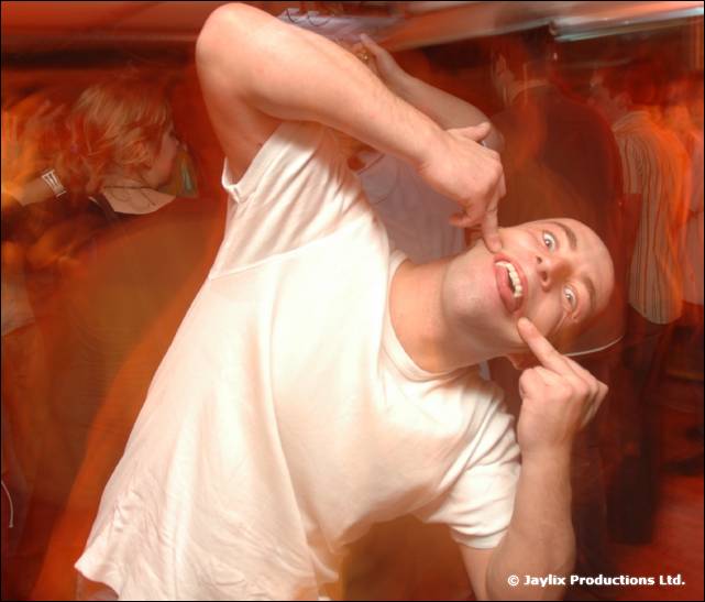 A bald person in a white T-shirt pulls a face, fingers pressed to their cheek, in a busy club