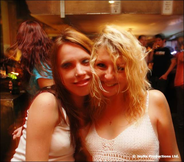 Two smiling women pose cheek to cheek in a bar, one with long dark hair and one with curly blonde hair