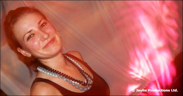 A smiling person with red hair and bead necklaces against a pink blurred background