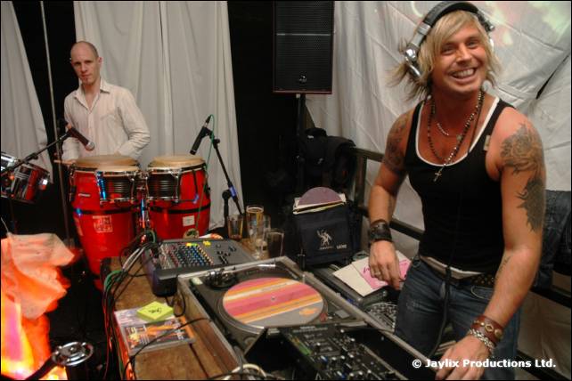 A smiling DJ in headphones and a black vest at the decks, with conga drums and a person behind