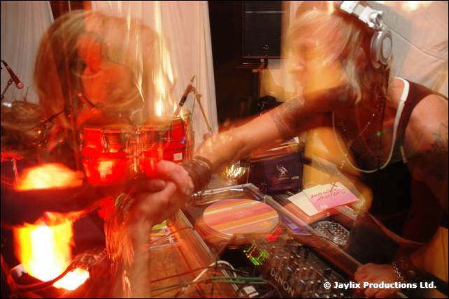 A DJ in headphones reaches across the decks beside red conga drums, with heavy motion blur