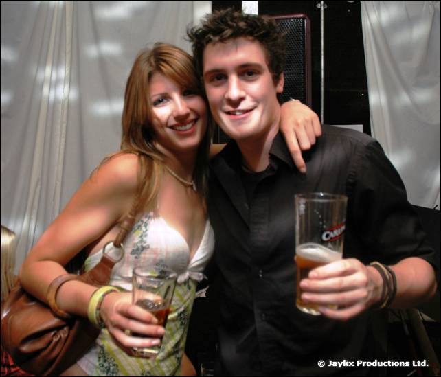 A smiling woman and a man in black pose with pints of beer in front of white drapes