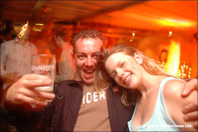 A man holding a Guinness glass and a woman with blonde hair smile at the camera in an orange-lit bar