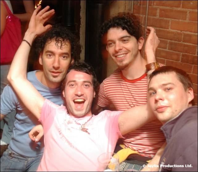 Five young men crowd together smiling, one in a pink T-shirt with an arm raised, in front of a brick wall