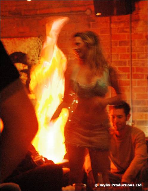 A large flame burns beside a person dancing in front of a brick wall, a seated person to the right