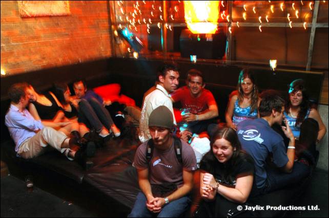 A group sits and stands on dark sofas in a bar under string lights, seen from above