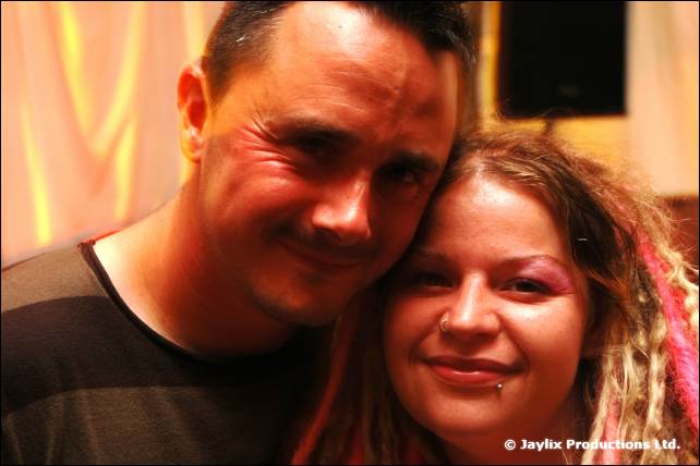 A smiling man and a woman with pink-streaked dreadlocks rest their heads together