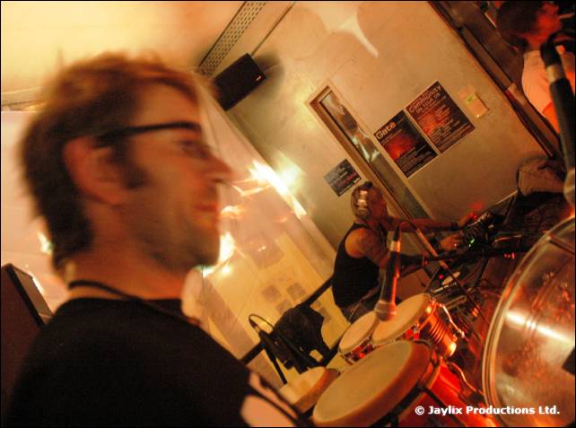 A person with glasses in the foreground beside a bongo player, with posters on the wall behind