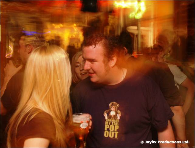 A man in a dark T-shirt holds a pint while a woman with long blonde hair stands in front of him, in a crowd