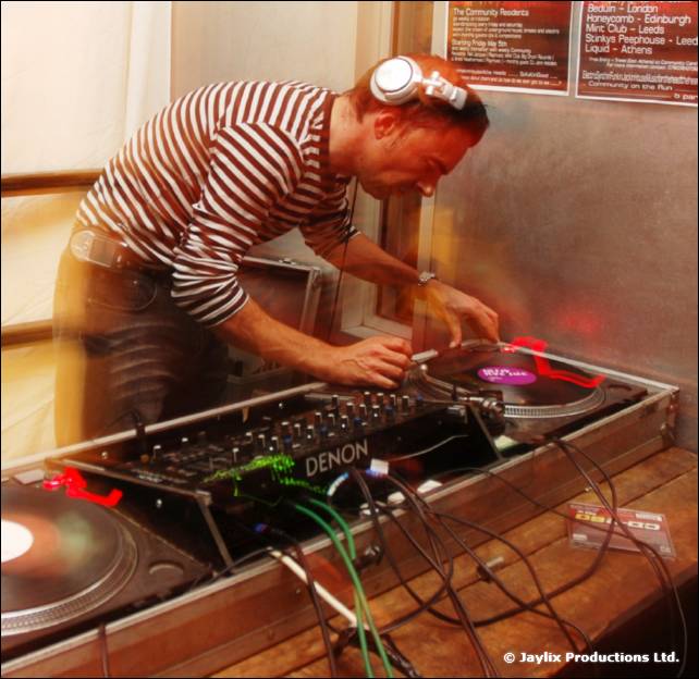 A DJ in striped shirt and headphones mixes at Denon mixer and turntables, with a flyer on the wall