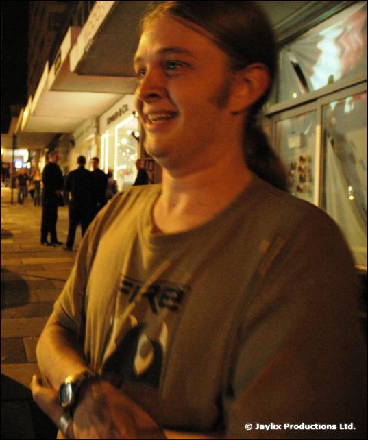 A smiling person with a ponytail in an olive T-shirt stands on a street at night