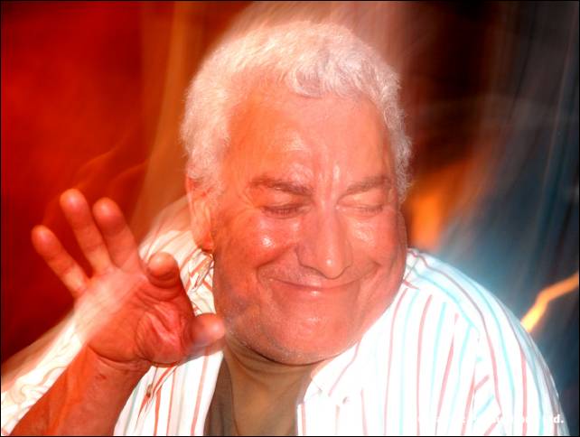 An older person with white hair and a striped shirt smiles with eyes closed, one hand raised