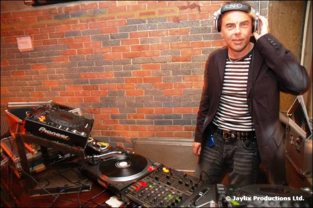 A DJ in a striped shirt and headphones stands beside Pioneer decks in front of a brick wall