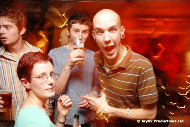 Four people pose in a bar, one bald person pulling a wide-eyed face, another holding a pint