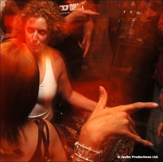 Dancers crowd together, a woman pointing a hand towards the camera, in a dark, orange-lit blur