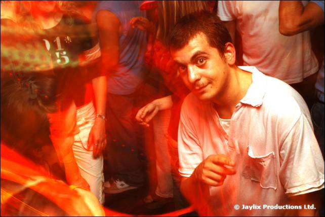 A man in a white polo shirt looks at the camera among dancing people in a blurred crowd