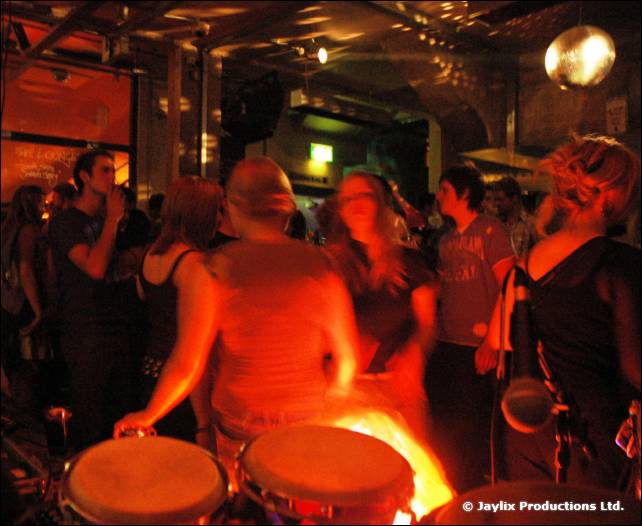 Bongo drums in the foreground and a crowd dancing in a bar with a mirrored ball above