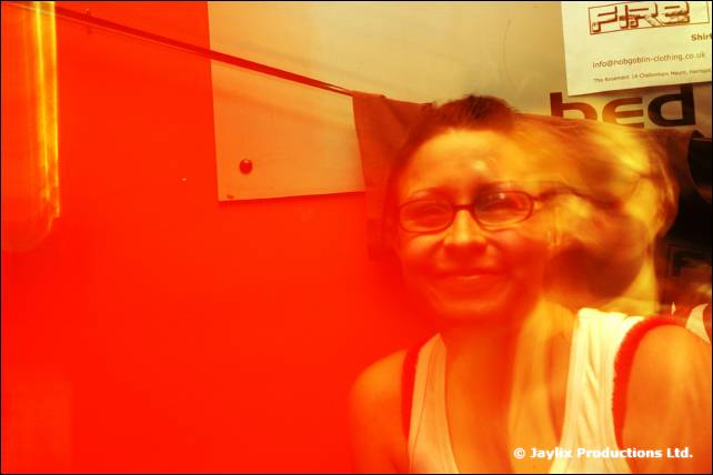 A smiling person with glasses in a red-lit room with a FIRE sign on the wall behind