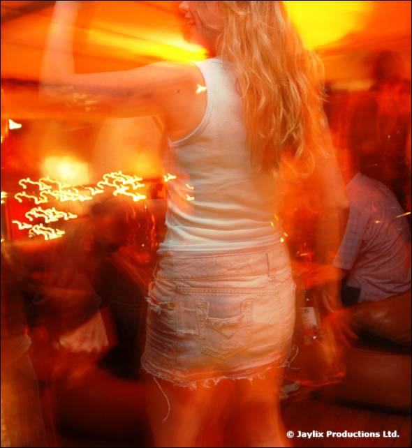 A person with long blonde hair in a white top and denim skirt dances, with orange light trails and blur