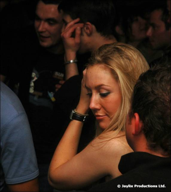 A woman with blonde hair looks down in a dark crowd, a man behind her touching his face