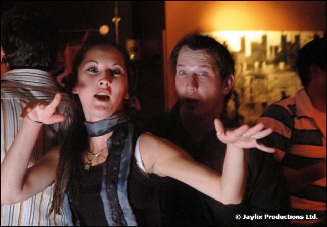 A woman in a scarf and a man behind her pull expressive faces in a bar, hands raised