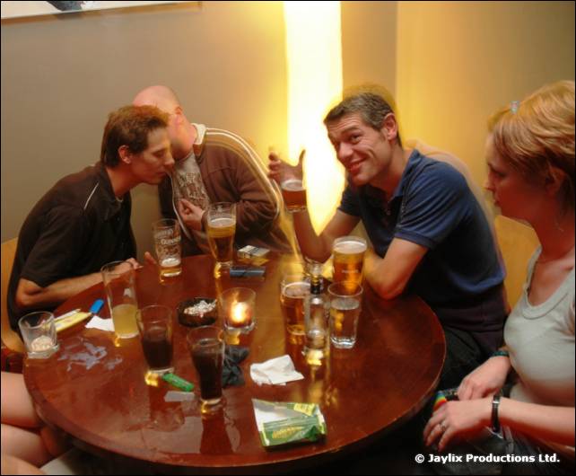 A group sits at a round table with pints and drinks in a warmly lit bar, two leaning close together