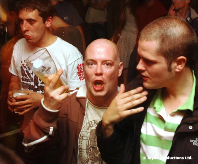Three men talk and gesture in a busy bar, one holding a glass, one bald in a brown jacket