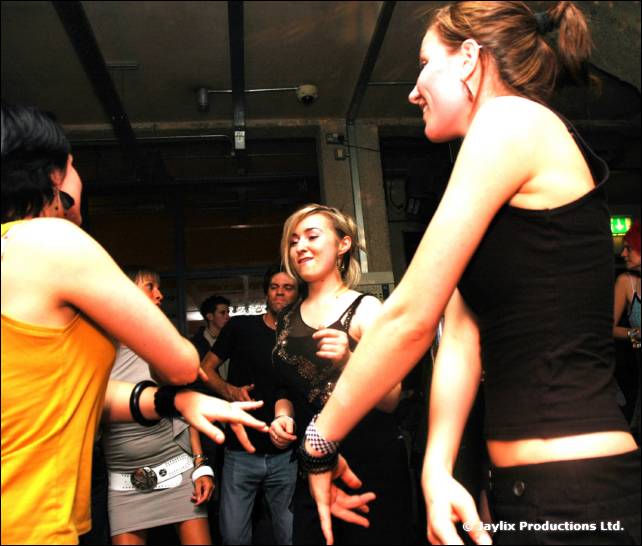 Dancers in a dark bar, including a person in a yellow top and a woman in black, reaching out