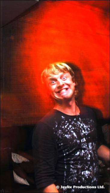 A person in a black patterned top grins with a wide-eyed expression against a red wall