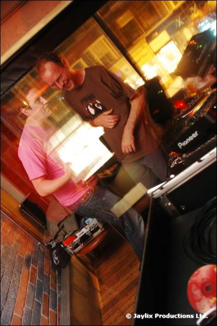 A person in a pink top and another in a brown T-shirt stand by Pioneer DJ equipment, seen at a tilt