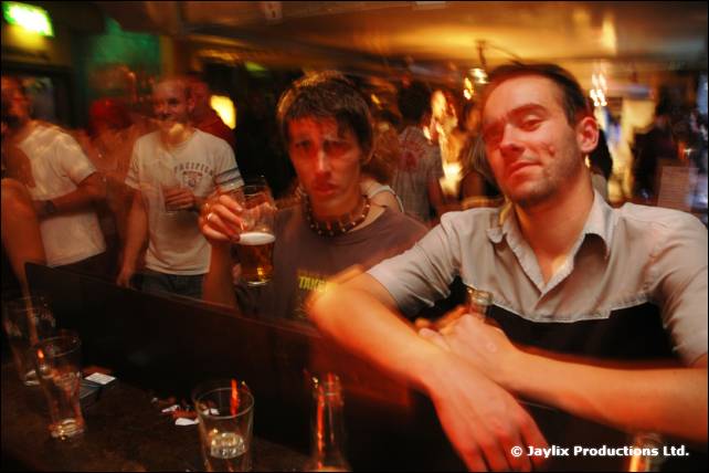 Two men lean on a bar, one holding a pint, with others behind