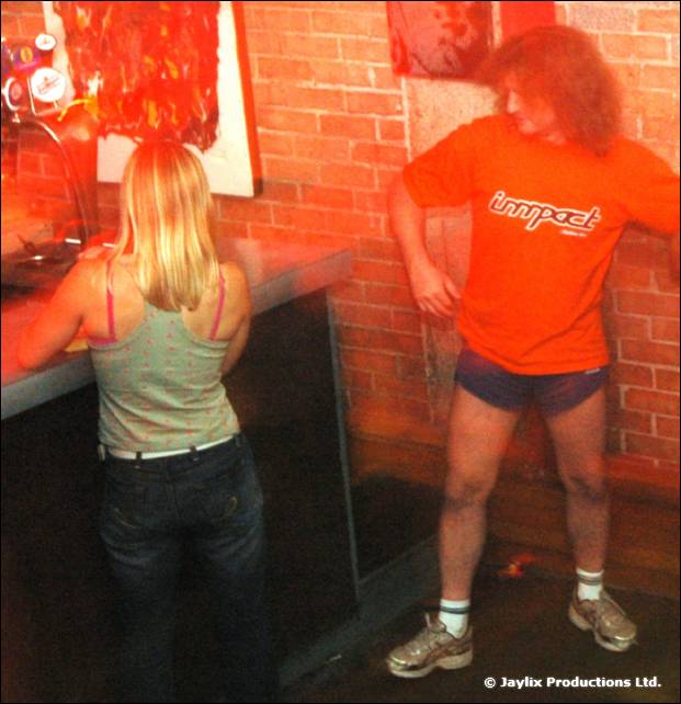 A woman at a bar and a person in an orange Impact T-shirt and shorts dance against a brick wall