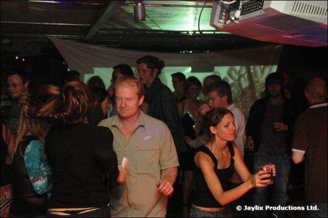 A crowd stands and dances in a dimly lit club, with a projector above
