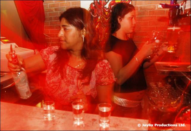 Two women serve behind a bar with shot glasses on the counter, one holding a bottle
