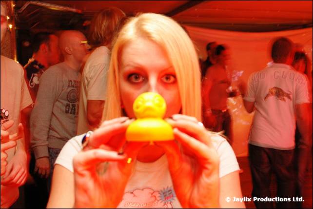 A woman with blonde hair holds a yellow rubber duck in front of her face in a red-lit bar