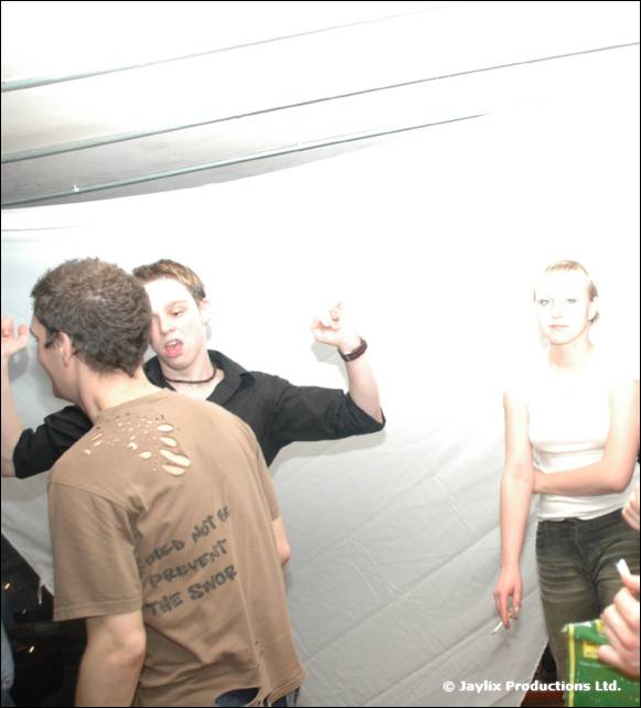 People dance against a white backdrop, one in a brown T-shirt with printed text, one smoking